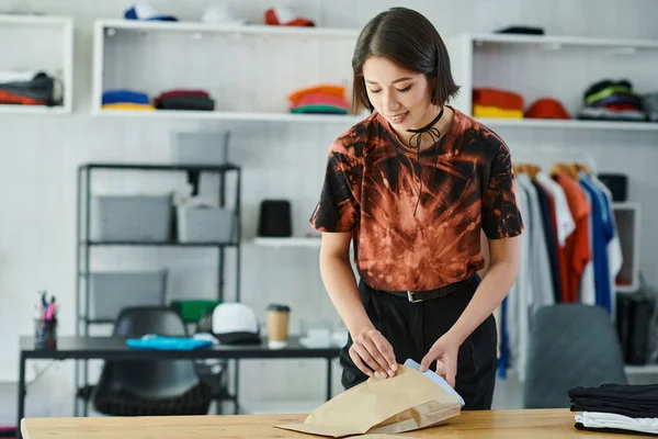Mujer asiática feliz embalaje cuidadosamente doblado prendas en la mesa en el estudio de impresión textil, industria de la moda - foto de stock