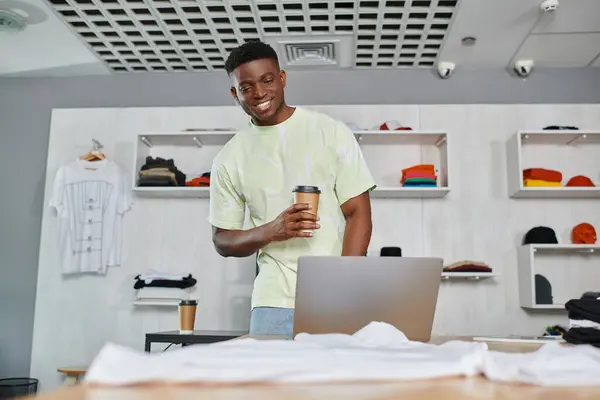 Sonriente diseñador afroamericano con café para ir a buscar portátil cerca de la ropa en el estudio de impresión - foto de stock