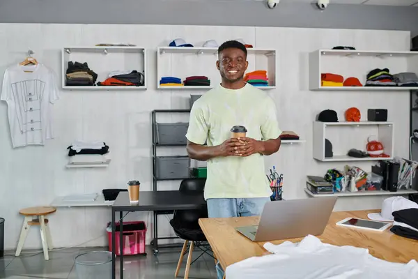 Designer americano africano feliz com café para ir olhando para a câmera perto do laptop no atelier impresso — Fotografia de Stock