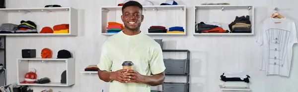 Alegre diseñador afroamericano con taza de papel cerca de estantes con ropa en el estudio de impresión, pancarta - foto de stock