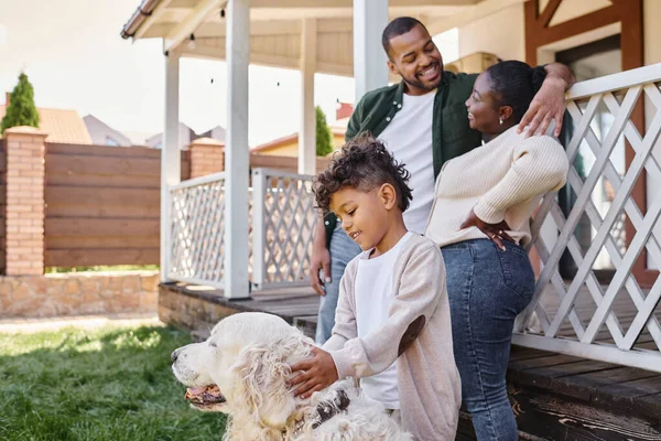 Allegro bambino afroamericano che gioca con il cane vicino ai genitori sul cortile della loro casa, tempo di qualità — Foto stock
