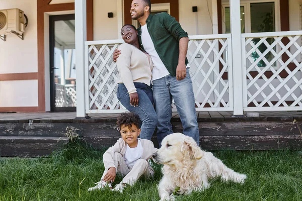 Familienzeit, glückliche afrikanisch-amerikanische Eltern umarmen sich, während Sohn mit Hund im Hinterhof des Hauses spielt — Stockfoto