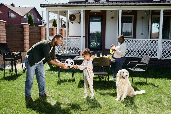 Семейный уик-энд, счастливый африканский американский отец дает футбольный мяч сыну рядом с женой накрывая стол — стоковое фото