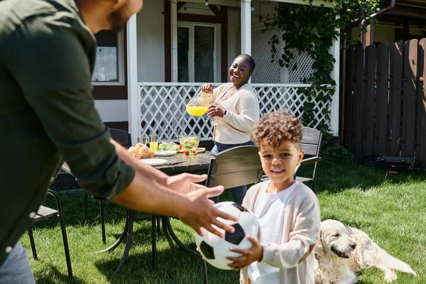 Happy african american woman smiling while holding jug with orange juice near family in house garden — Stock Photo