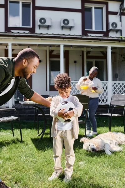 Famiglia tempo, felice padre afroamericano guardando il figlio con pallone da calcio vicino moglie sul cortile — Foto stock