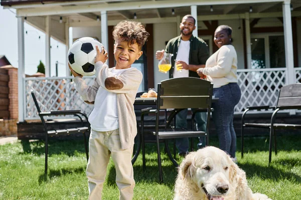 Tempo de família, garoto americano africano alegre segurando futebol perto de pais e cachorro no quintal da casa — Fotografia de Stock