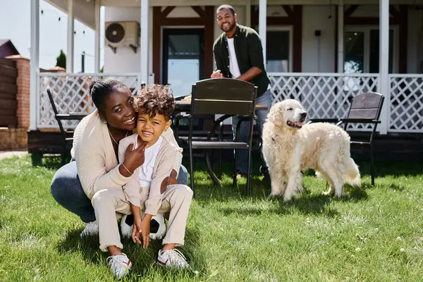 Alegre africano americano madre abrazando hijo sentado en fútbol cerca marido en patio trasero de casa — Stock Photo