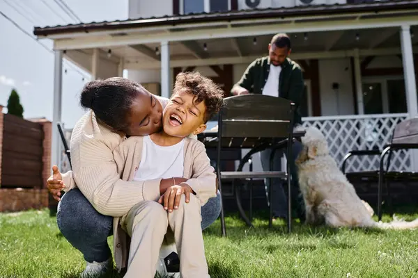 African american mother kissing cheek of excited son near husband and family dog on backyard — Stock Photo