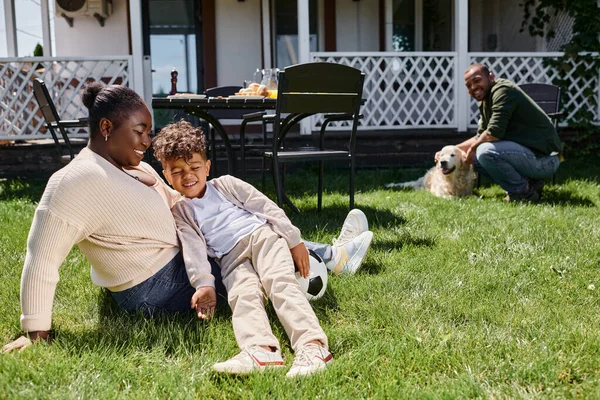 Cheerful african american mother sitting near son next to football near husband on backyard of house — Stock Photo