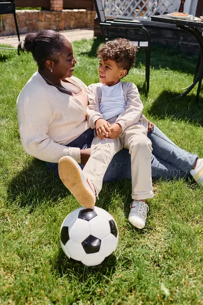 Happy african american mother sitting on lawn with son next to soccer ball on backyard of house — Stock Photo