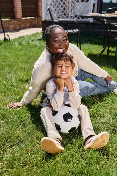 Cheerful african american mother sitting on lawn with son next to soccer ball on backyard of house — Stock Photo