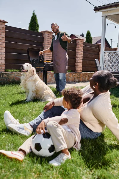 African american mother sitting on lawn with son near dog while happy husband cooking on bbq grill — Stock Photo