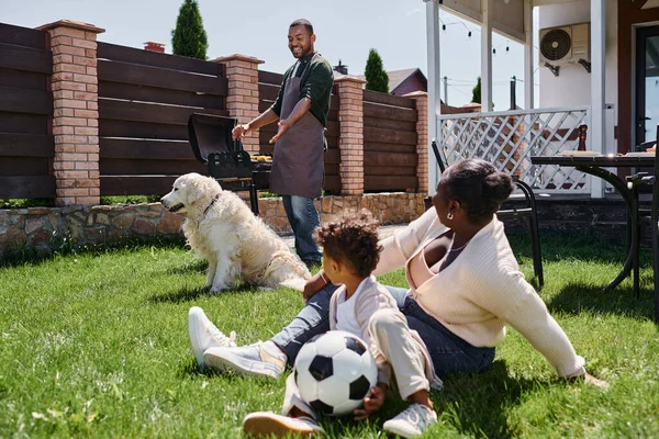 Afrikanisch-amerikanische Mutter sitzt mit Sohn neben Hund auf Rasen, während positiver Mann auf Grill kocht — Stockfoto