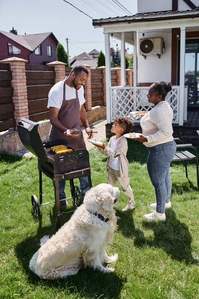 Happy african american man cooking grilled corn on bbq grill near dog, wife and son on backyard — Stock Photo