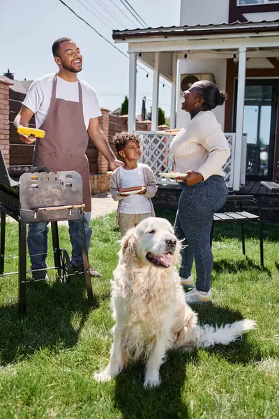 Веселый африканский американец готовит кукурузу на гриле возле собаки, жены и сына на заднем дворе — стоковое фото