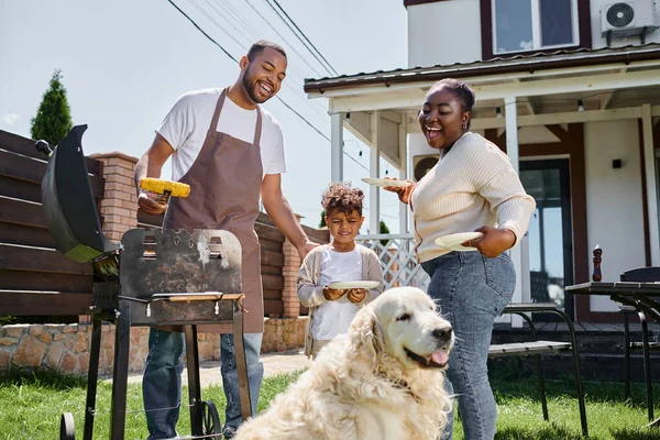 Feliz homem americano africano preparando milho grelhado na churrasqueira perto do cão, esposa e filho no quintal — Fotografia de Stock