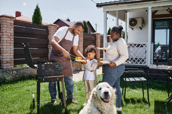 Hombre afroamericano feliz sirviendo maíz a la parrilla en el plato de hijo cerca de esposa y perro en el patio trasero - foto de stock