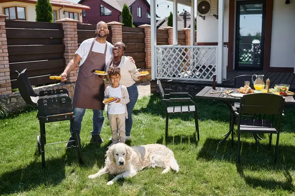Positivo afro-americano família segurando placas com milho grelhado perto churrasqueira e cão no quintal — Fotografia de Stock