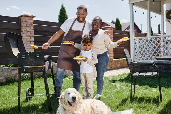 Happy african american man cooking grilled corn on bbq grill near wife and son and looking at dog — Stock Photo