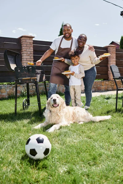 Família africana feliz segurando pratos com milho grelhado perto churrasqueira e cão no quintal — Fotografia de Stock