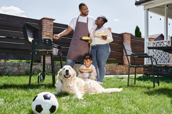 Familia afroamericana alegre celebración de platos con maíz a la parrilla cerca de barbacoa parrilla y perro en el patio trasero - foto de stock