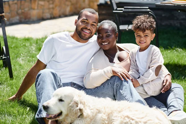 Retrato familiar de la familia afroamericana positiva mirando a la cámara y sentado en el césped cerca del perro - foto de stock