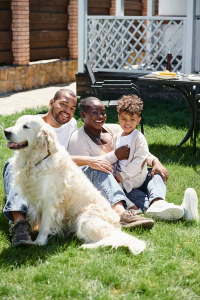 Family portrait of joyful african american parents and son smiling and sitting on lawn near dog — Stock Photo