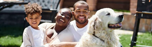 Bandera de la familia de alegres padres afroamericanos e hijo sonriendo y sentado en el césped cerca del perro - foto de stock