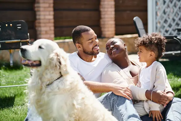 Сімейний час радісних афроамериканських батьків і сина, посміхаючись і сидячи на траві біля собаки — стокове фото