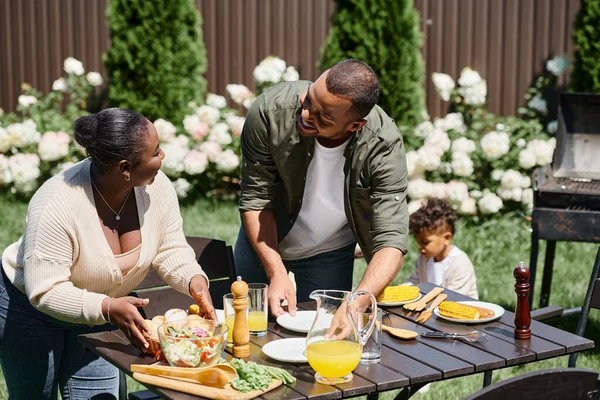 Alegre afroamericano padres sirviendo mesa en jardín cerca hijo jugando en patio trasero, familia - foto de stock
