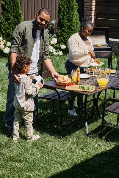 Весёлые африканские американские родители, обслуживающие столик в саду рядом с сыном, держащим футбольный мяч на заднем дворе — стоковое фото