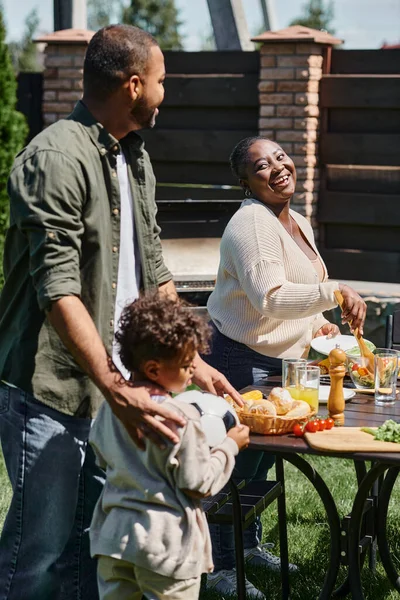 Padres afroamericanos felices sirviendo comida en la mesa en el jardín cerca de hijo sosteniendo pelota de fútbol - foto de stock