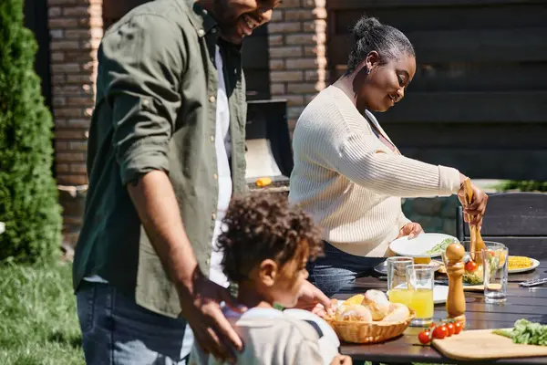 Fröhlich afrikanisch-amerikanische Eltern servieren Essen auf Tisch im Garten in der Nähe Sohn hält Fußball — Stockfoto
