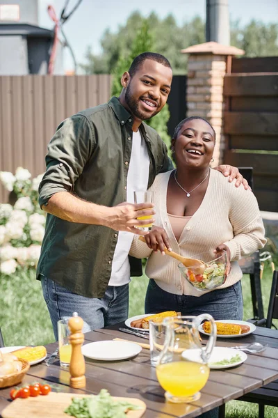 Happy african american man hugging wife mixing salad in glass bowl while having bbq on backyard — Stock Photo