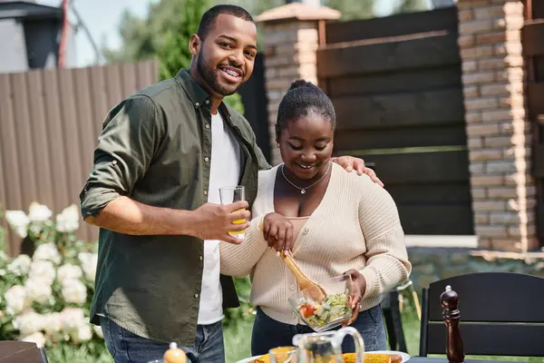 Веселый африканский американец обнимает жену, смешивая салат в стеклянную миску, пока ест барбекю на заднем дворе — стоковое фото