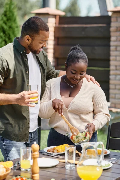 Happy african american man hugging wife holding salad in glass bowl while having bbq on backyard — Stock Photo