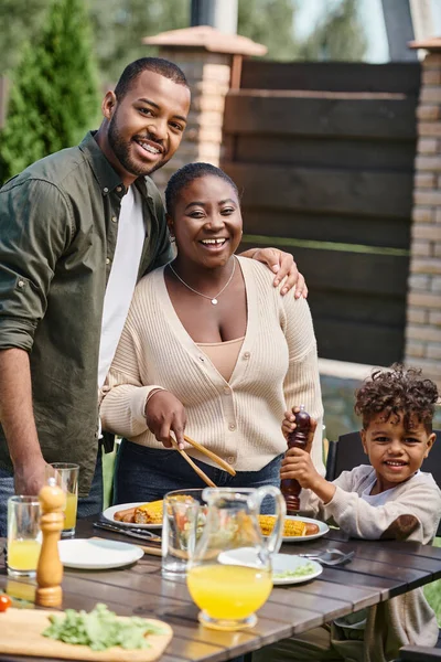 Retrato de pais afro-americanos felizes e filho preparando mesa de jardim para almoço no quintal — Fotografia de Stock