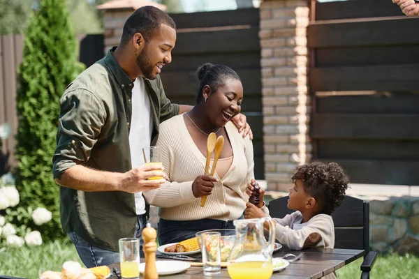 Retrato de padres afroamericanos felices y su hijo preparando la mesa para el almuerzo en el jardín en el patio trasero - foto de stock