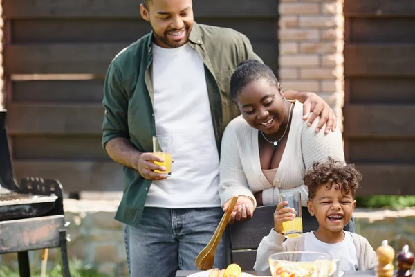 Африканские американские родители смотрят на радостного сына, держащего стакан апельсинового сока на заднем дворе во время барбекю — стоковое фото
