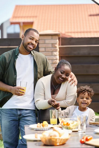Retrato de padres afroamericanos felices e hijo preparando mesa de jardín para el almuerzo en el patio trasero - foto de stock