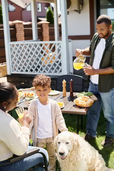 Happy african american boy smiling and standing near dog and parents during family bbq on backyard — Stock Photo