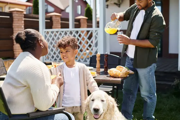 Joyeux garçon afro-américain souriant et debout près du chien et des parents pendant le barbecue familial sur la cour — Photo de stock