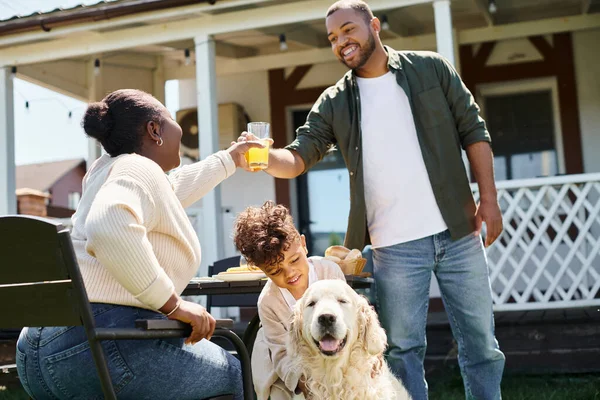 Веселый африканский американский мальчик улыбается и ласкает собаку рядом с родителями во время семейного барбекю на заднем дворе — стоковое фото