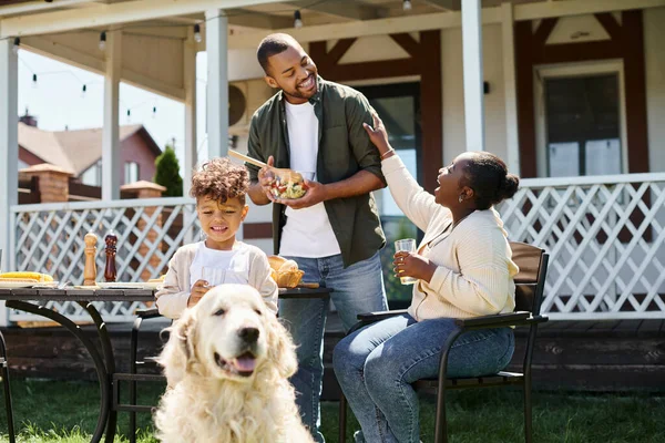 Dog looking at camera and sitting on green lawn near african american family during bbq on backyard — Stock Photo