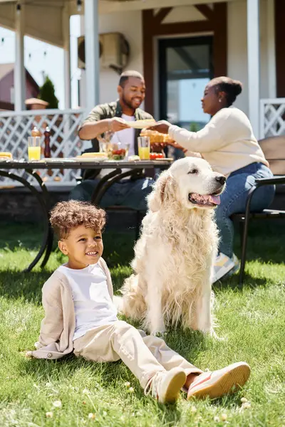 Alegre menino americano africano sentado no gramado verde perto do cão e os pais tendo refeição no jardim — Fotografia de Stock