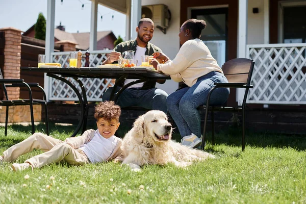 Happy african american kid sitting on green lawn near dog and parents having lunch in garden — Stock Photo