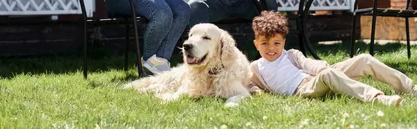 Happy african american kid sitting on green lawn near dog and parents on backyard, banner — Stock Photo