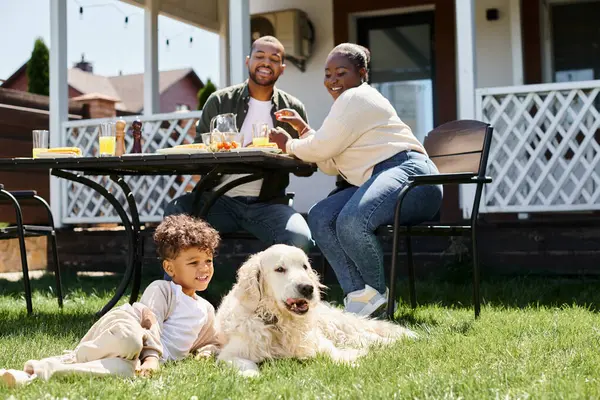 Garoto americano africano positivo sentado no gramado verde perto do cão e pais almoçando no jardim — Fotografia de Stock