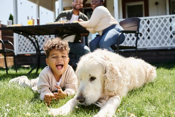 Niño afroamericano sorprendido sentado en el césped verde cerca del perro y los padres almorzando en el jardín - foto de stock