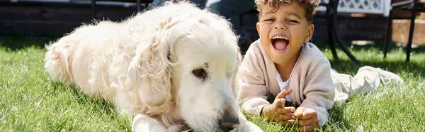 Amazed african american boy lying on green lawn next to family dog on backyard, horizontal banner — Stock Photo